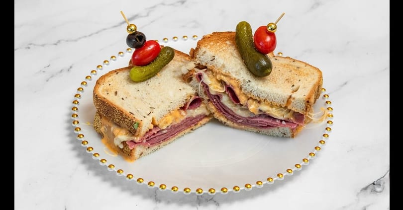 A toasted pastrami sandwich, cut in half, with melted cheese and sauce, garnished with pickles and tomatoes.