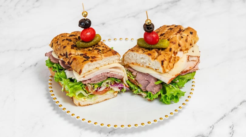 Two halves of a deli sandwich with roast beef, turkey, cheese, and vegetables on focaccia bread.
