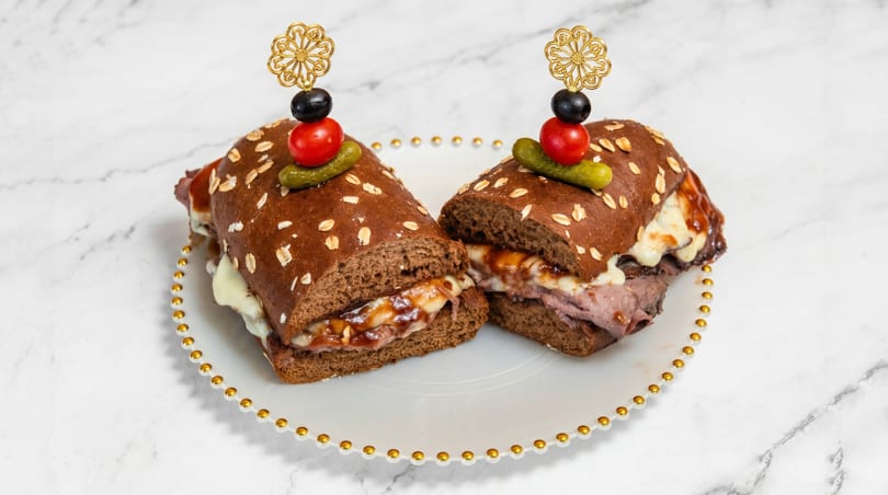 Two roast beef sandwiches with melted cheese and sauce on oat-topped bread, garnished with olives, tomato, and pickle.