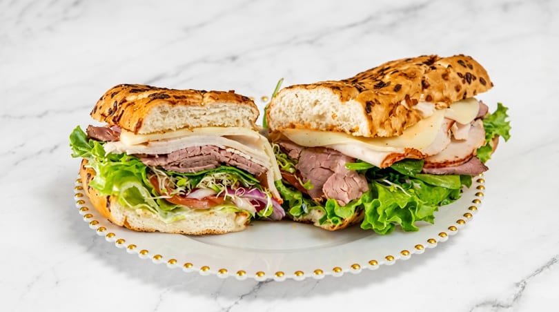 A large deli sandwich, cut in half, filled with meats, cheese, lettuce, and sprouts on a white plate.